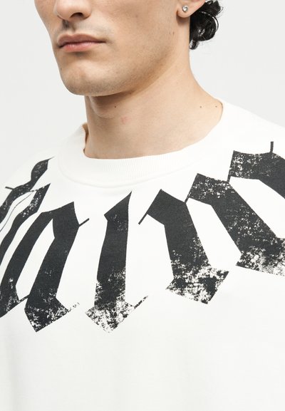 Κοντινό πλάνο ενός ατόμου που φορά λευκό πουκάμισο με μεγάλο, φθαρμένο μαύρο γοτθικό κείμενο στο στήθος και ένα μικρό εμφανές καρφί στο αυτί.