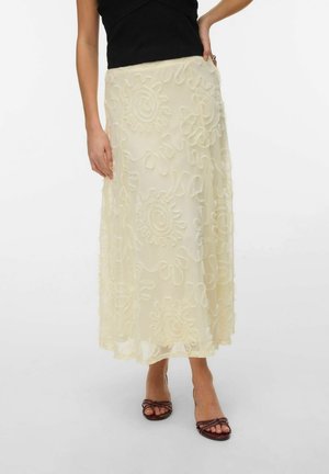 Žena nosi krémovú midi sukňu s textúrovanými vírivými vzormi, v kombinácii s čiernym tielkom bez rukávov a hnedými otvorenými topánkami na podpätku.