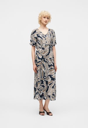 Donna con capelli biondi ricci che indossa un vestito midi con fantasia a foglie blu navy, beige e bianco, con maniche a palloncino e sandali neri con tacco e punta aperta.