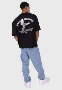 Svart oversized t-skjorte med et grafisk motiv av en hvit due og tekst på ryggen; kombinert med lyseblå, baggy jeans og hvite sneakers.