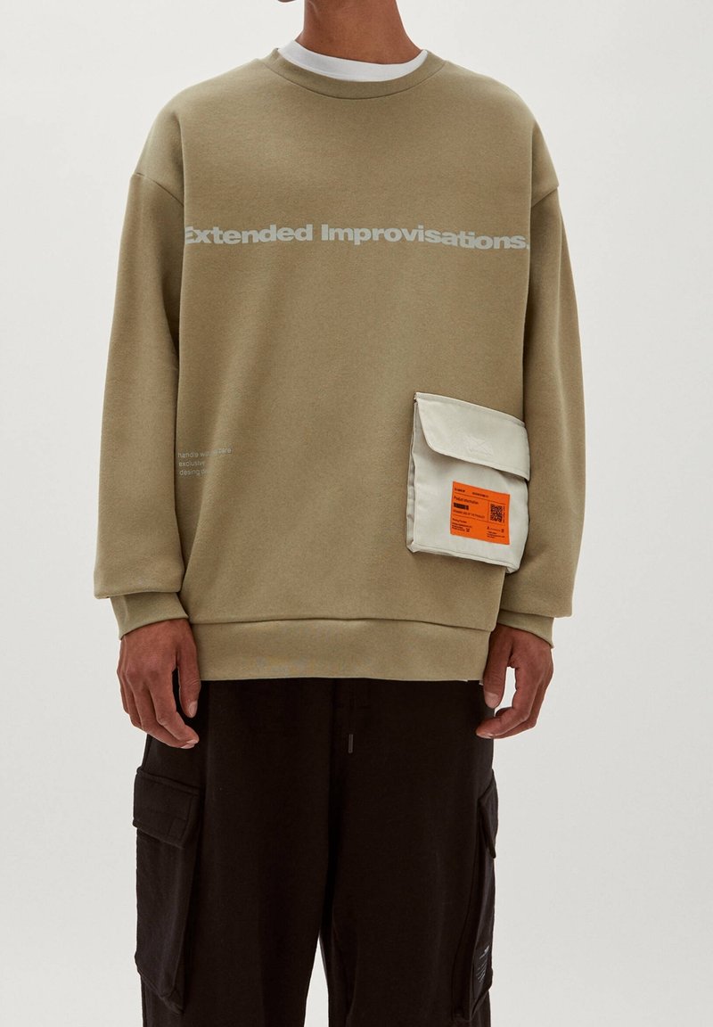 Sweat-shirt beige avec le texte "Extended Improvisations", poche poitrine blanche avec étiquette orange, porté avec un pantalon cargo noir.