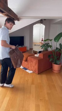 Brun lærveske, rektangulær form, jevn tekstur; ligger på et tregulv ved siden av en oransje kordfløyelssofa og en potteplante.