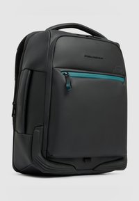 Mochila negra elegante con asa acolchada, bolsillo frontal con cremallera color verde azulado, base redondeada reforzada y logotipo de la marca sutil en superficie lisa.