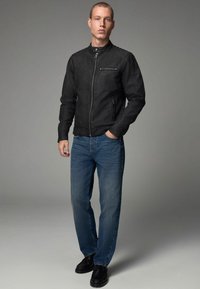Chaqueta de cuero negra con cuello alzado, cremallera frontal y dos bolsillos laterales. Llevada sobre jeans azules y zapatos negros. Textura suave.