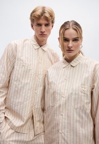 Δύο μοντέλα φοράνε μπεζ πουκάμισα με κουμπιά και κάθετες ρίγες σε ανοιχτό μπλε χρώμα. Κάθε πουκάμισο έχει τσέπες με πατς και κλασικό γιακά.