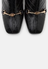 Zapatos de cuero negro con un patrón acolchado, punta afilada y acentos de metal dorado. Presentan correas con eslabones circulares y un cierre decorativo.
