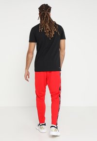 Homme avec de longues dreadlocks portant un t-shirt noir, un pantalon de sport rouge avec des rayures sur les côtés et des baskets blanches, se tenant de dos.
