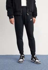 Černé Nike tepláky s elastickým pasem, zúženými nohavicemi a vyšitým logem. V kombinaci s černo-bílými Nike teniskami. Texturovaná tkanina.