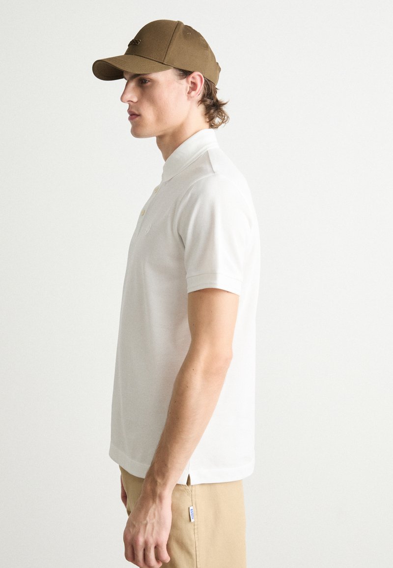 Bela polo majica s kratkimi rokavi, ovratnikom in zapenjanjem na gumb, v kombinaciji z rjavim šistrom. Slim fit oblikovanje, gladka tekstura, casual stil.