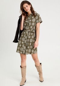 Robe verte à manches courtes avec un motif floral blanc, ceinturée à la taille. Portée avec une veste noire et des bottes de cowboy beiges.
