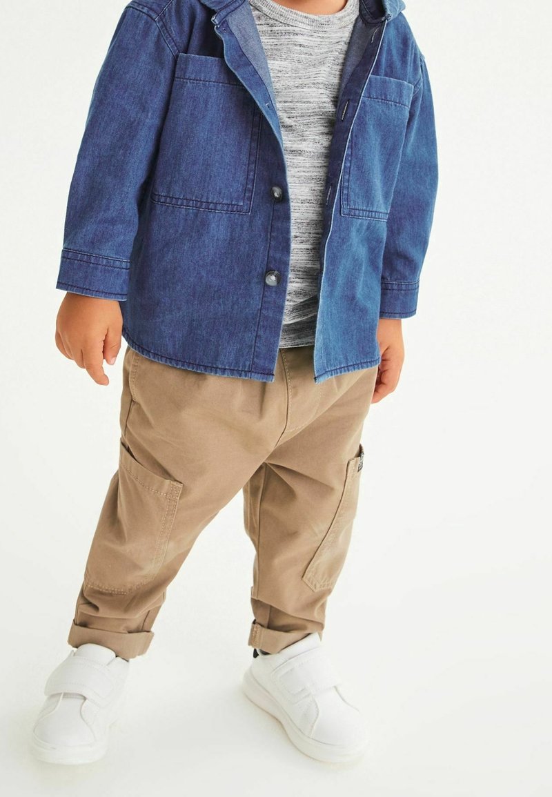 Enfant portant une veste en denim bleu, une chemise grise rayée, un pantalon cargo beige et des baskets blanches à scratch, debout sur un fond clair.
