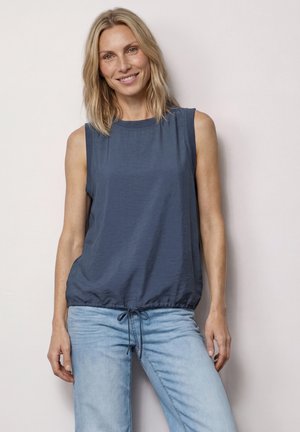 Lachende vrouw met schouderlang blond haar die een mouwloze blauwe top met koord in de taille en lichtblauwe spijkerbroek draagt, staand tegen een lichte muur.