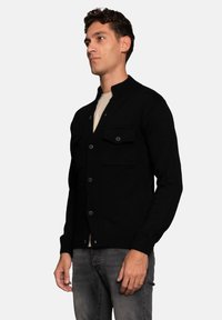 Black knitted cardigan with a high collar, featuring two chest pockets and button closure. Worn over a light-colored shirt and paired with dark jeans.