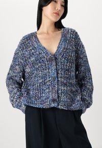 Strikket cardigan i blå og lilla nyanser, med V-hals, lange ermer og store knapper. Strukturert, avslappet passform med en chunky design.