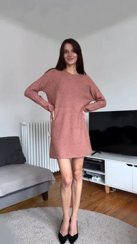 Robe en maille côtelée rose poudré avec manches longues et motif géométrique. Longueur courte, portée avec des talons hauts noirs sur un sol en bois.
