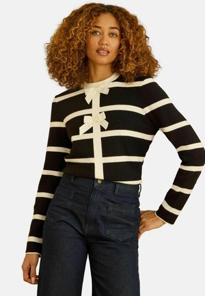 Schwarz-weiß gestreifter Pullover mit rundem Ausschnitt, vorne mit zwei Schleifen, aus weichem Material. Kombiniert mit dunklen Denim-Jeans.