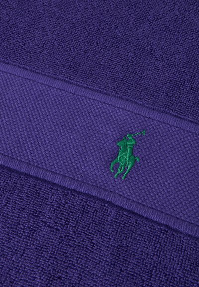 Μωβ υφασμάτινη υφή με πράσινο κεντημένο λογότυπο παίκτη πόλο στο κέντρο μιας υφασμένης οριζόντιας ταινίας.