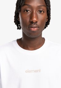 Camiseta de algodón blanca con cuello redondo y un logotipo minimalista en beige "element" en el frente. Textura suave y diseño de ajuste estándar.