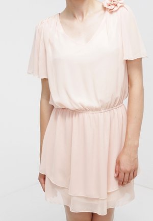 Femme portant une robe en chiffon rose clair à manches courtes superposée, avec un détail fleur sur l'épaule et une taille cintrée.
