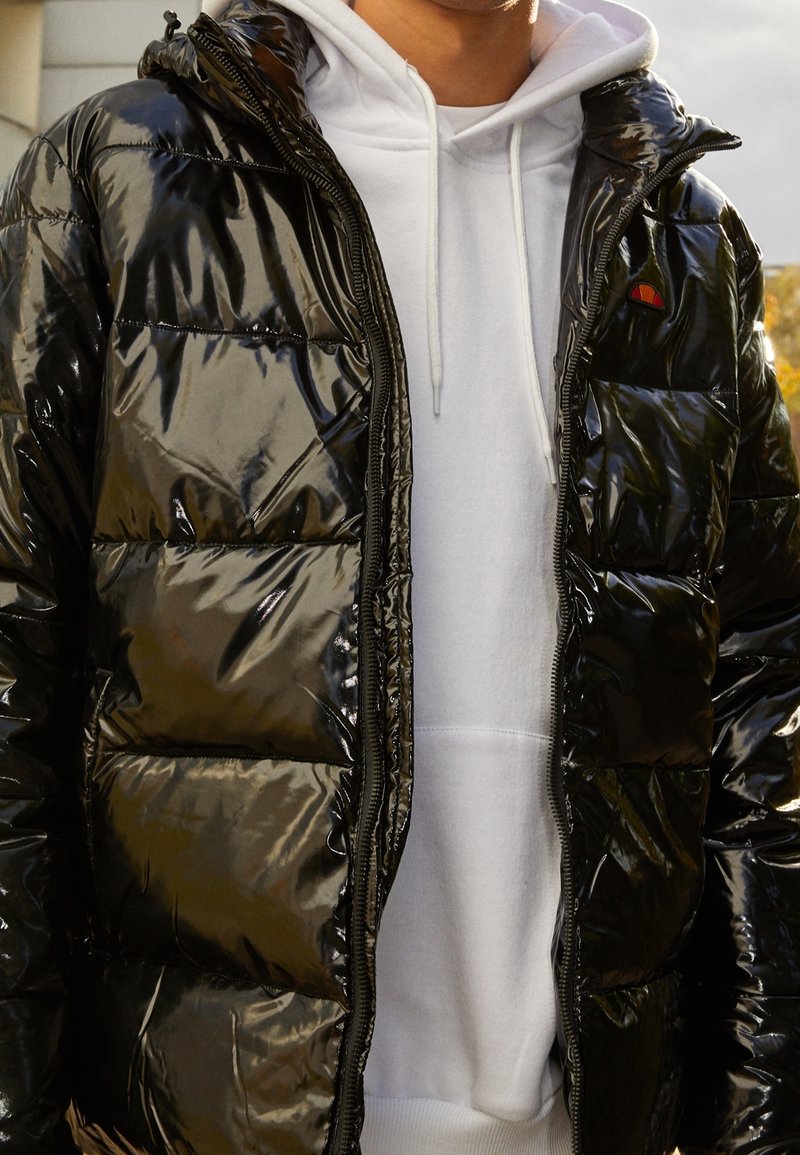 Černá lesklá prošívaná bunda na zip s kapucí a bočními kapsami. Nošena přes bílou mikinu s kapucí, zvýrazňující kontrastní barvy.