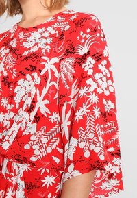 Femme portant un vêtement rouge avec des motifs tropicaux de fleurs et de feuilles en blanc et noir, détails de manches amples, et boucle d'oreille visible.