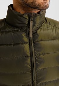 Olivgrüne, gefütterte Jacke mit hohem Kragen und Reißverschluss. Verfügt über gesteppten Abschnitte und eine glatte, glänzende Textur.