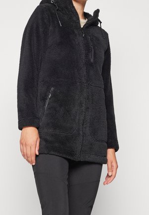 Personne portant une veste polaire noire avec capuche, fermeture éclair frontale et poches zippées, associée à un pantalon noir.