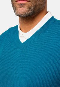 Pull en tricot turquoise avec un design à col en V, présentant une texture côtelée. Une chemise blanche à col est visible sous le pull.