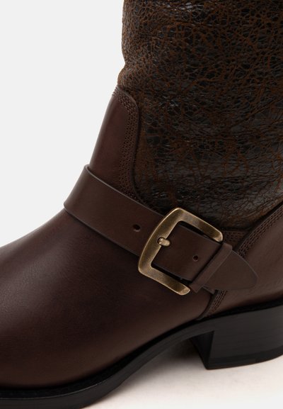 Bottine en cuir marron à cheville avec dessus texturé, bride décorative, boucle en laiton et petit talon noir sur fond blanc.