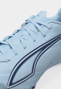 Šviesiai mėlynas futbolo batų modelis su tekstūruotu paviršiumi, šviesiai mėlynomis raišteliais ir tamsiai mėlynu kampuotu logotipu šone.