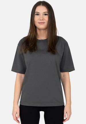 Jonge vrouw met steil bruin haar, die een eenvoudige losse donkergrijze korte mouwen t-shirt en zwarte broek draagt, staand tegen een lichte achtergrond.