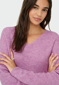 Jeune femme aux longs cheveux noirs portant un pull en tricot rose, les bras croisés, avec une bague en argent au doigt annulaires gauche.