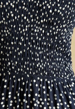 Marineblåt stof med hvide polkaprikker har smocket elastisk syning, der skaber en rynket tekstur på kjolens midtersektion.