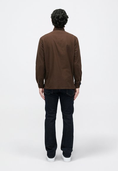 Camisa de mangas longas castanha, em mistura de algodão, com colarinho e punhos abotoados, usada com jeans escuros e ténis brancos. Vista traseira visível.