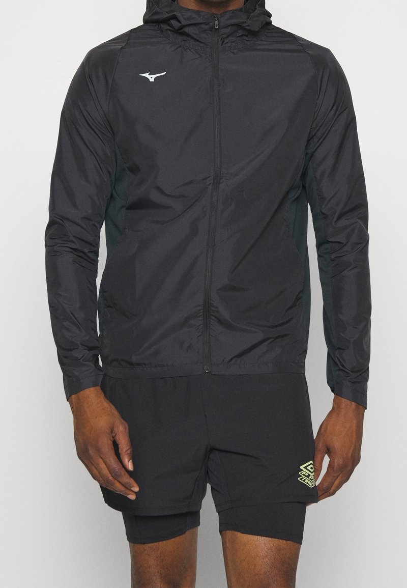 Homme portant une veste à capuche noire zippée et un short de sport noir avec logo sur la cuisse gauche, debout devant un fond uni.