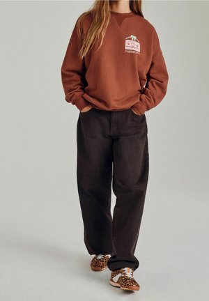 Junge Person geht und trägt einen rostfarbenen Pullover mit Wintergrafik, schwarze weiter Hose und Leopardenmuster-Sneaker. Hände in den Taschen.