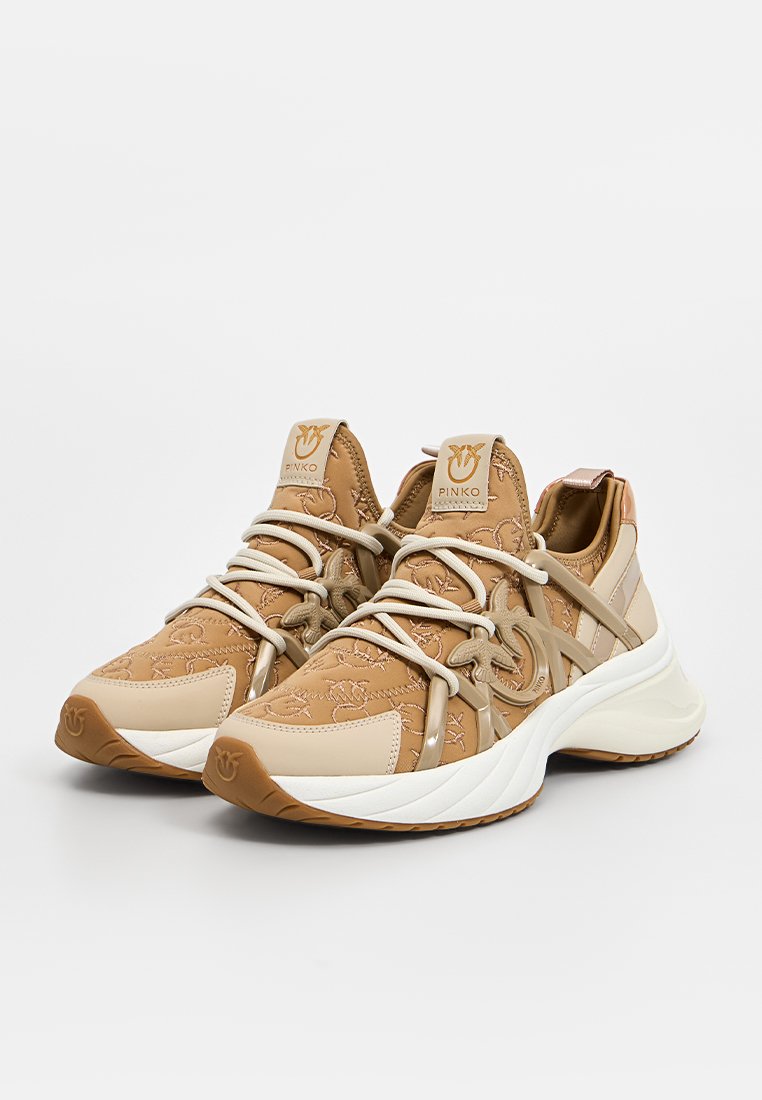 Baskets de sport beige avec tissu texturé, coutures décoratives et lacets élastiques. Présentent une semelle intermédiaire blanche et une semelle en caoutchouc beige.