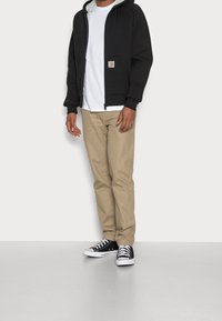 Uomo che indossa una felpa nera con zip, una maglietta bianca, pantaloni beige e scarpe da ginnastica nere basse, in piedi contro uno sfondo chiaro uniforme.