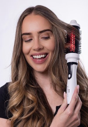 Der Haarbürsten-Föhn mit einem weißen Griff, schwarzen Borsten und rotem Licht. Lange, lockige Haare werden frisiert, vor einem schlichten Hintergrund.