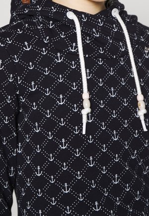 Sweat à capuche bleu marine avec ancre blanche et motif losange en pointillés, avec cordons blancs ornés de perles roses et beiges.
