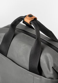 Grijze tas met zwarte stoffen handvatten en een tan leren accent. Beschikt over een ritssluiting en zichtbare stiksels aan de zijkant. Gladde textuur.