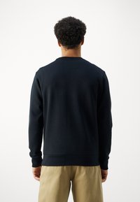 Hombre con cabello rizado, vistiendo una camisa de manga larga negra y pantalones beige, de espaldas frente a un fondo blanco liso.