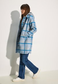 Cappotto in misto lana con motivo a righe blu e grigie, dotato di cappuccio, bottoni e tasche frontali, abbinato a jeans blu e sneakers bianche.