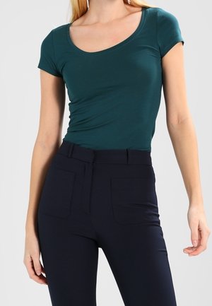 Femme portant un haut moulant vert foncé à col rond et manches courtes, ainsi qu'un pantalon noir taille haute avec poches avant, devant un fond uni.