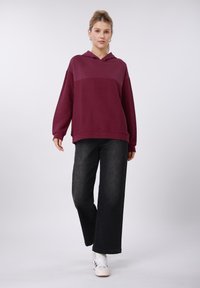 Sweat à capuche bordeaux avec motif texturé, manches larges et poche avant, associé à un jean noir ample et des baskets blanches.