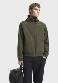 Olivenfarbene Softshell-Jacke mit Kapuze, Reißverschluss, elastischen Bündchen und Taschen. Trägt man zu schwarzen Cargo-Hosen und einem schwarzen Rucksack.