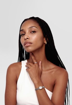 Donna con lunghe trecce che indossa un top bianco a una spalla, toccandosi il collo, con indosso un orologio d'argento, orecchini e anelli.