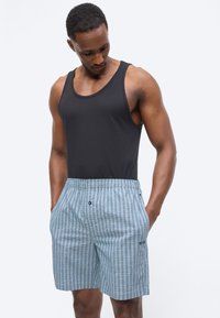 Homme musclé portant un débardeur noir et un short à carreaux bleus, les mains dans les poches, regardant vers le bas.