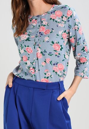 Haut floral bleu clair avec des roses roses et de petits accents, avec un col boutonné et des manches trois-quarts. Associé à un pantalon bleu.