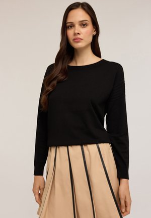 Maglione lungo nero a maniche lunghe con collo rotondo e dettagli a costine sulle spalle, abbinato a una gonna beige plissettata con strisce nere.